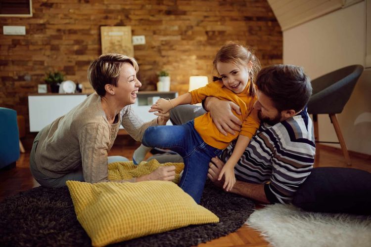 Happy family of three playing on the living room floor. Mom and dad laugh while tickling their cheerful daughter.