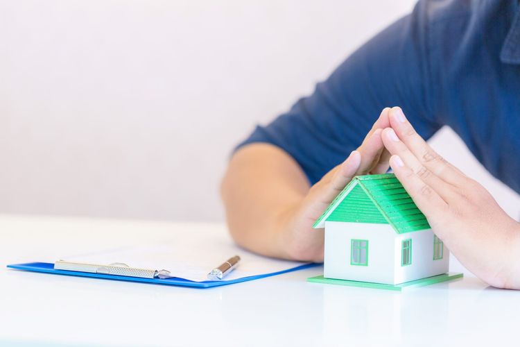 Person's hands cupping a small white model house with a green roof on a table next to a clipboard and pen.