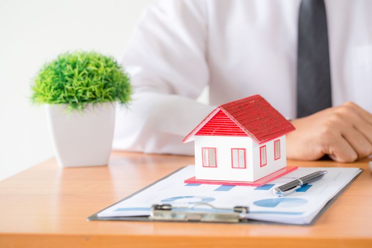 Small house model on business documents with a pen, plant, and a person in shirt and tie at a desk.