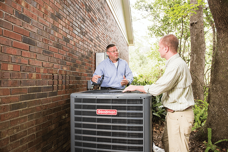 HVAC technician explaining an outdoor Goodman AC unit to a homeowner next to a brick wall and trees.