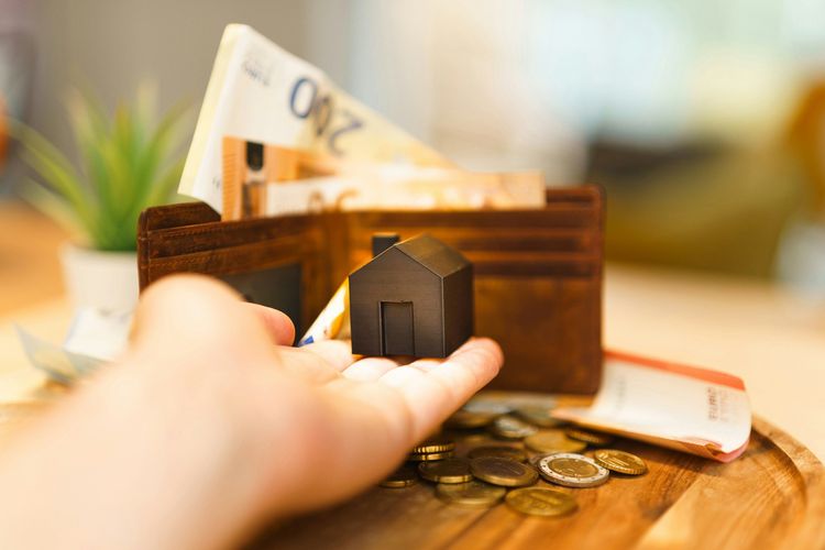 Hand holding a miniature black house model with a wallet, euro banknotes, and coins in the background.