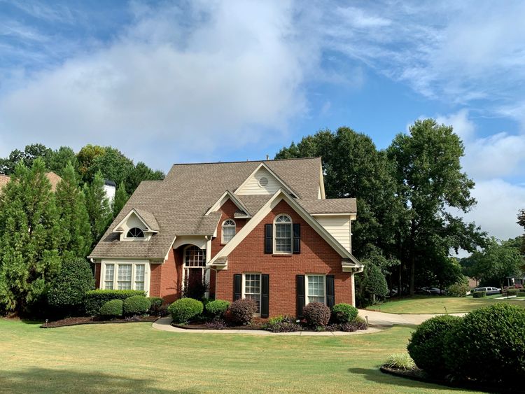 Large red brick house with light trim, gray shingle roof, green lawn, shrubs, and trees under a blue sky.
