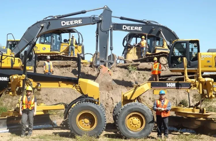 Construction workers in safety gear pose with multiple John Deere excavators, bulldozers, and graders on a dirt worksite.