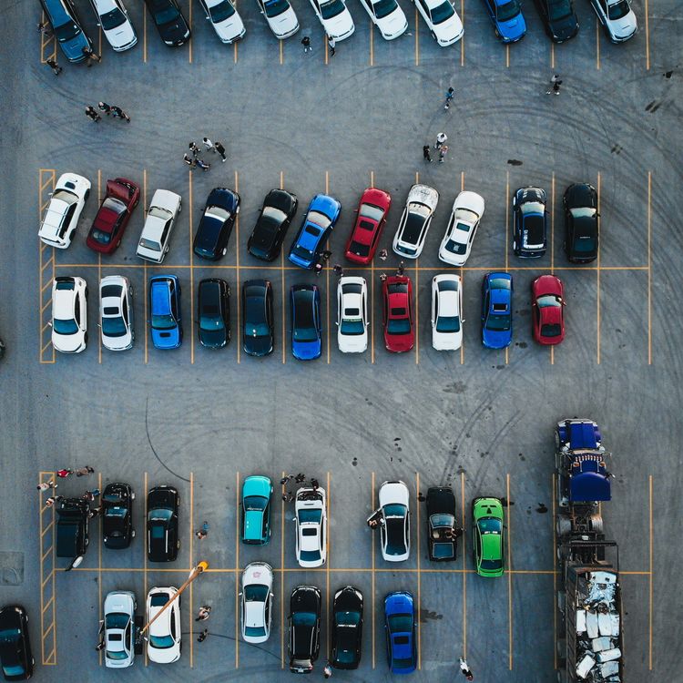 Overhead view of a bustling parking lot with many colorful cars, groups of people, and a truck full of scrap metal.