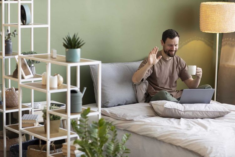 Smiling man on bed waves during a video call on laptop, holding a mug. Bedroom with green wall and decorative shelf.