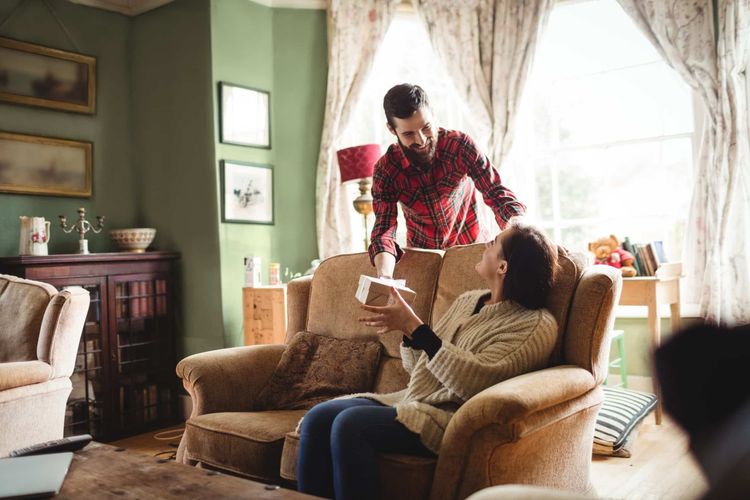 Man in plaid shirt gives a gift to a smiling woman sitting on a couch in a cozy living room.