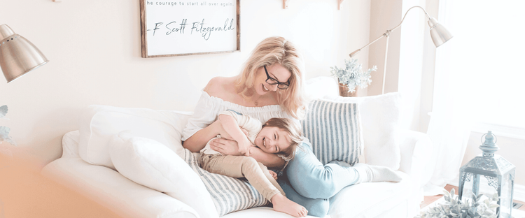 A blonde woman in glasses smiles while cuddling and tickling her laughing child on a white sofa with striped pillows.