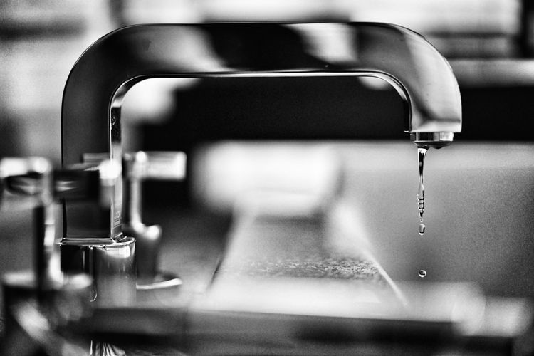 Monochrome close-up of a shiny faucet, with several water droplets forming a chain as they drip from the spout.