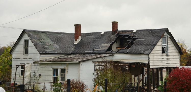 Dilapidated white house with a severely damaged roof, peeling shingles, and structural collapse under a cloudy sky.