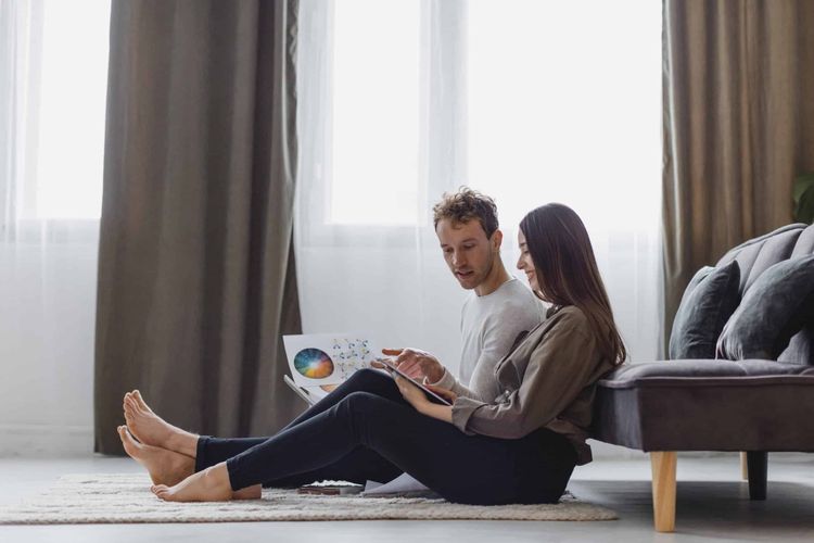 Couple sitting on rug at home, woman pointing at tablet as man looks at documents with colorful charts.