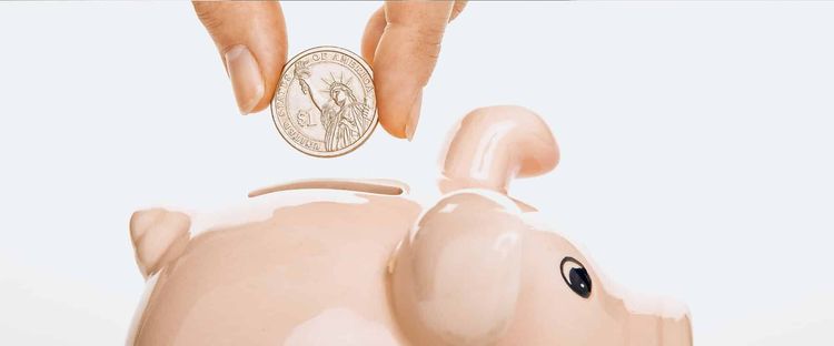 Hand inserting a $1 coin into the slot of a light pink piggy bank.