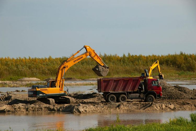 Yellow Hyundai excavator loads sand into a red dump truck next to water, with tall grasses in the background.
