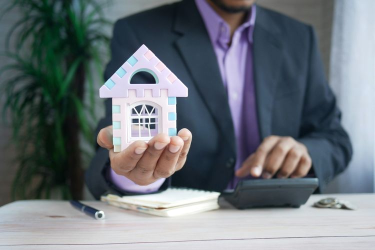 Person in suit holds a pastel model house while using a calculator, with a notebook and coins on the table.