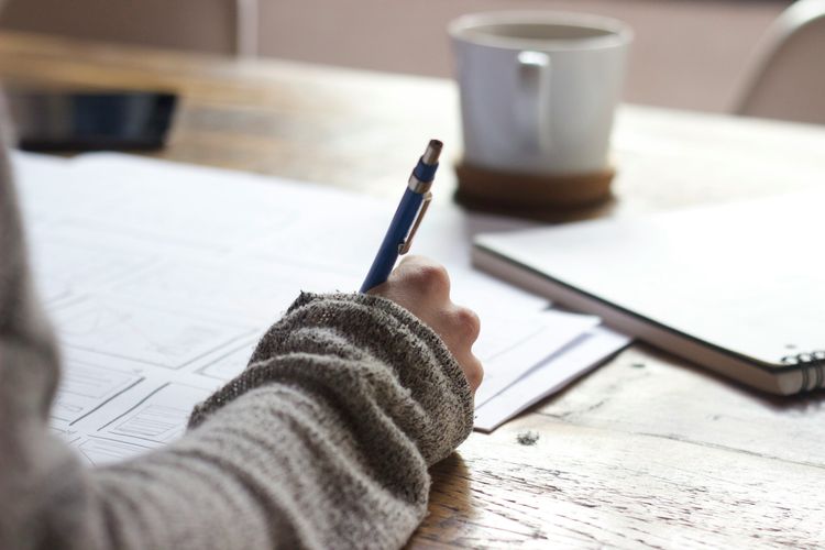 Close-up of a hand drawing UI sketches on paper with a pen on a wooden desk. A coffee mug is in the background.