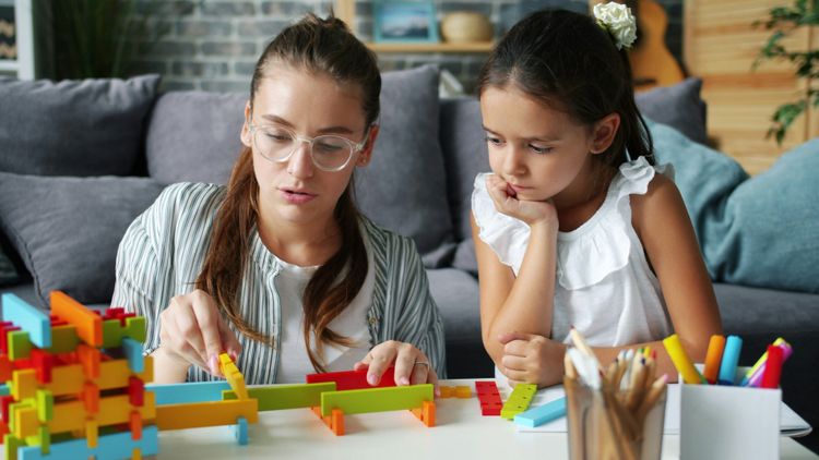 A woman and young girl build a colorful structure with interlocking blocks. The woman places a piece, the girl watches intently.