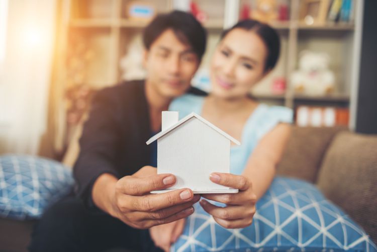Blurred smiling couple jointly holds a white miniature house model, centered and in focus.