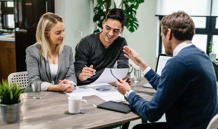 Three people in a bright office meeting. A man gestures, explaining charts to a smiling man and woman across the table.