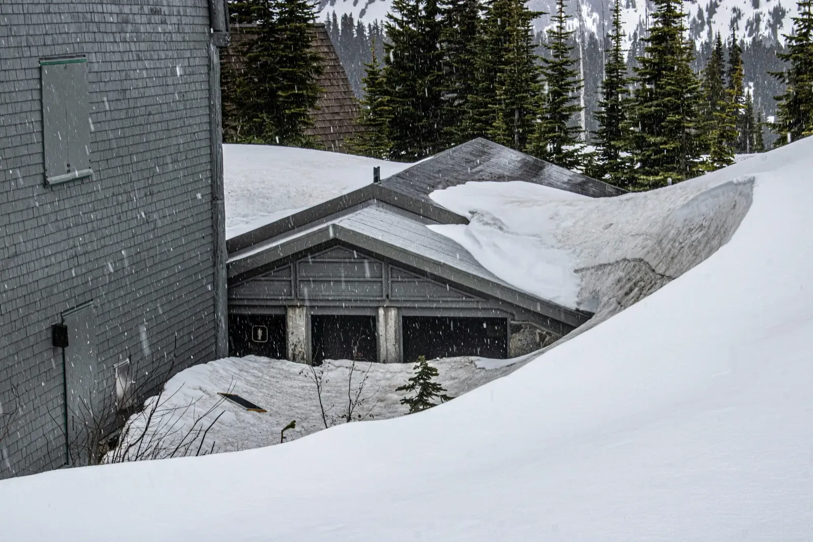 Snowing heavily on grey shingled buildings with deep snowdrifts, pine trees, and snow-covered mountains in the background.