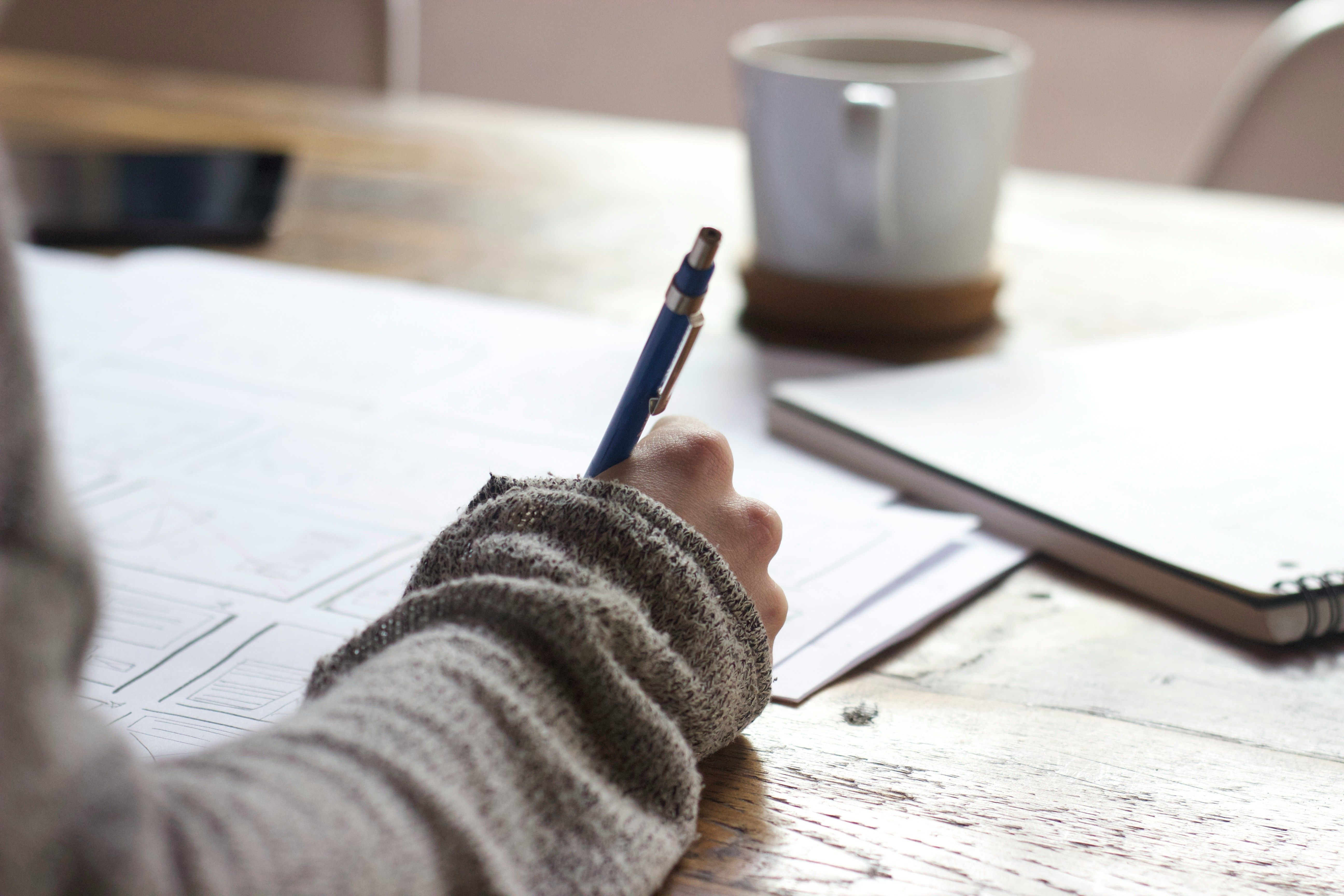 Close-up of a hand drawing UI sketches on paper with a pen on a wooden desk. A coffee mug is in the background.